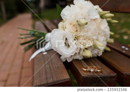 White Wedding Bouquet with Gold Rings on Wooden Bench in Garden Setting 135931586