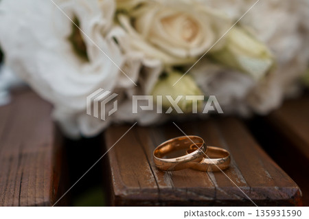 Elegant Gold Wedding Rings on Wooden Surface with White Rose Bouquet in Soft Focus Background Elegant Gold Wedding Rings on Wooden Surface with White Rose Bouquet in Soft Focus Background 135931590