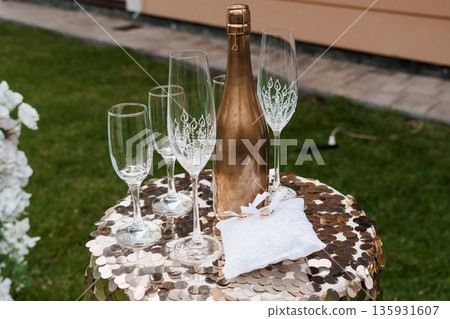 Elegant Gold Champagne Bottle and Crystal Glasses on Sequined Table with White Floral Arrangement Outdoors Elegant Gold Champagne Bottle and Crystal Glasses on Sequined Table with White Floral Arrangement Outdoors 135931607