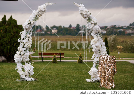 Elegant White Floral Wedding Arch with Gold Sequin Podium in Outdoor Garden Setting Under Overcast Sky Elegant White Floral Wedding Arch with Gold Sequin Podium in Outdoor Garden Setting Under Overcast Sky 135931608