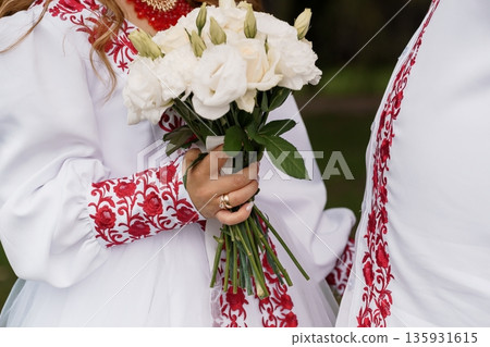 Traditional Ukrainian Wedding Dress with Red Embroidery and White Rose Bouquet in Natural Outdoor Setting Traditional Ukrainian Wedding Dress with Red Embroidery and White Rose Bouquet in Natural Outdoor Setting 135931615