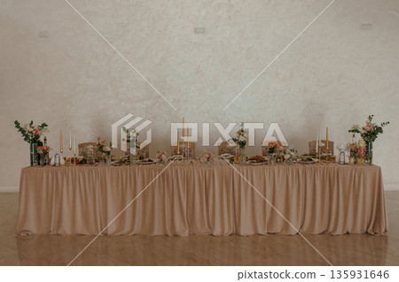 Elegant Long Banquet Table with Gold Candles and Floral Arrangements in Soft Beige Linen Setting Elegant Long Banquet Table with Gold Candles and Floral Arrangements in Soft Beige Linen Setting 135931646