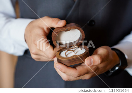 Elegant Wooden Ring Box Holding Two Gold Wedding Bands Held by Groom in Formal Suit 135931664
