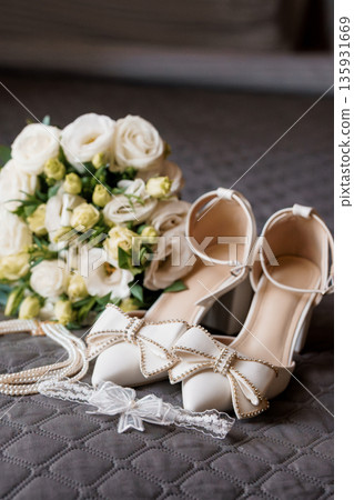 Elegant White Wedding Bouquet with Pearl-Embellished Bridal Shoes and Garter on Quilted Gray Background Elegant White Wedding Bouquet with Pearl-Embellished Bridal Shoes and Garter on Quilted Gray Background 135931669