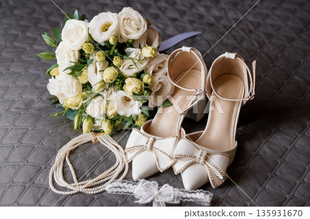 Elegant White Wedding Bouquet with Pearl Necklace and Bow-Adorned Bridal Shoes on Quilted Gray Background Elegant White Wedding Bouquet with Pearl Necklace and Bow-Adorned Bridal Shoes on Quilted Gray Background 135931670