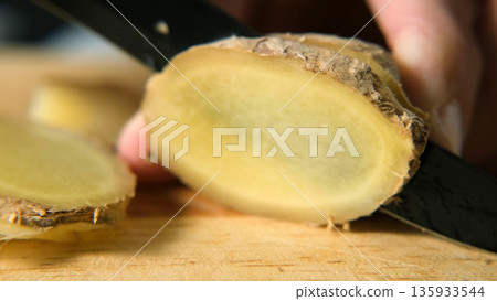 Macro of slicing fresh ginger root on a wooden surface, showing texture and freshness. Natural detox and healthy lifestyle concept Macro of slicing fresh ginger root on a wooden surface, showing texture and freshness. Natural detox and healthy lifestyle concept 135933544