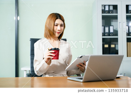 Confident beautiful Asian businesswoman typing laptop computer and digital tablet while holding coffee 135933718