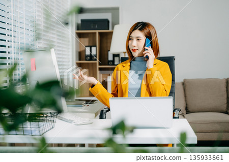 Confident Asian woman in yellow blazer using smartphone while working at desk with laptop and desktop in bright office. Modern, smart, and professional. Confident Asian woman in yellow blazer using smartphone while working at desk with laptop and desktop in bright office. Modern, smart, and professional. 135933861