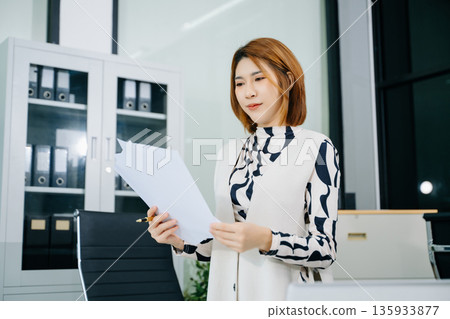 Young Asian woman in office analyzing paper reports, planning strategy with laptop and documents. Young Asian woman in office analyzing paper reports, planning strategy with laptop and documents. 135933877