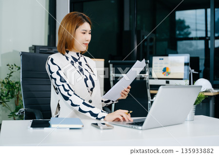 Young beautiful woman typing on tablet and laptop while sitting at the working white table in office 135933882