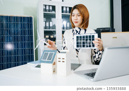 Asian businesswoman in office holding green globe with solar house and wind turbine models symbolizing renewable energy, sustainability Asian businesswoman in office holding green globe with solar house and wind turbine models symbolizing renewable energy, sustainability 135933888