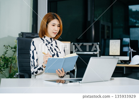 Asian businesswoman writing in notebook at modern office desk with laptop. A concept of planning, success 135933929