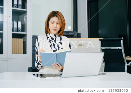 Asian businesswoman writing in notebook at modern office desk with laptop. A concept of planning, success Asian businesswoman writing in notebook at modern office desk with laptop. A concept of planning, success 135933930