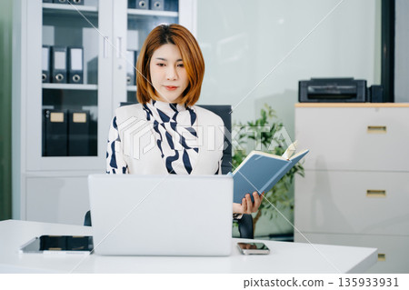 Asian businesswoman writing in notebook at modern office desk with laptop. A concept of planning, success Asian businesswoman writing in notebook at modern office desk with laptop. A concept of planning, success 135933931