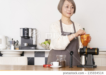 一位老婦人在廚房裡用咖啡研磨機研磨咖啡豆，並用義式咖啡機製作濃縮咖啡。 135934514