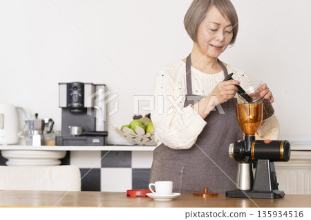 A senior woman grinding coffee beans in a coffee grinder and making espresso in an espresso machine in the kitchen A senior woman grinding coffee beans in a coffee grinder and making espresso in an espresso machine in the kitchen 135934516