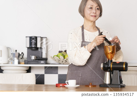 一位老婦人在廚房裡用咖啡研磨機研磨咖啡豆，並用義式咖啡機製作濃縮咖啡。 135934517