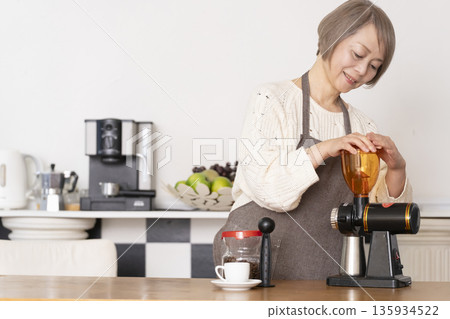 A senior woman grinding coffee beans in a coffee grinder and making espresso in an espresso machine in the kitchen 135934522