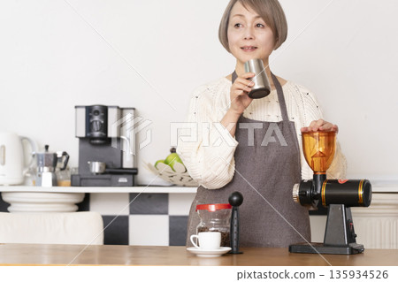 A senior woman grinding coffee beans in a coffee grinder and making espresso in an espresso machine in the kitchen A senior woman grinding coffee beans in a coffee grinder and making espresso in an espresso machine in the kitchen 135934526