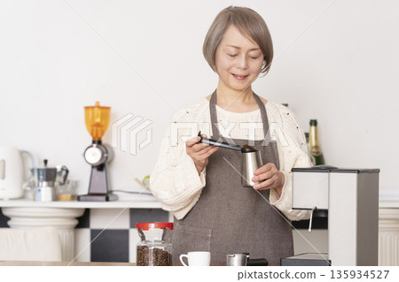 一位老婦人在廚房裡用咖啡研磨機研磨咖啡豆，並用義式咖啡機製作濃縮咖啡。 135934527