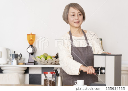 一位老婦人在廚房裡用咖啡研磨機研磨咖啡豆,並用義式咖啡機製作濃縮咖啡。 一位老婦人在廚房裡用咖啡研磨機研磨咖啡豆,並用義式咖啡機製作濃縮咖啡。 135934535