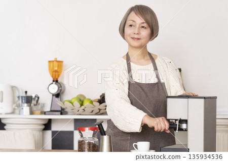 一位老婦人在廚房裡用咖啡研磨機研磨咖啡豆,並用義式咖啡機製作濃縮咖啡。 一位老婦人在廚房裡用咖啡研磨機研磨咖啡豆,並用義式咖啡機製作濃縮咖啡。 135934536