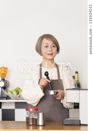 一位老婦人在廚房裡用咖啡研磨機研磨咖啡豆，並用義式咖啡機製作濃縮咖啡。 135934551