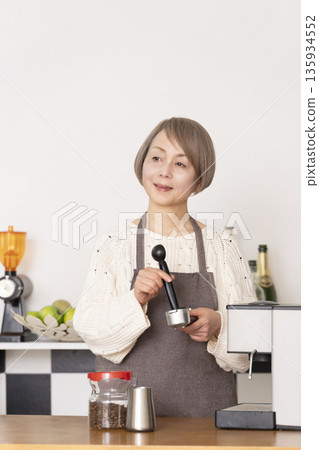 一位老婦人在廚房裡用咖啡研磨機研磨咖啡豆，並用義式咖啡機製作濃縮咖啡。 135934552