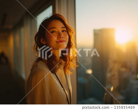 Smiling Japanese businesswoman looking at the city view in the office at dusk 135935489