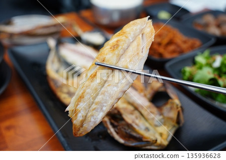 Chopsticks lifting a piece of crispy, golden grilled mackerel fillet at a Korean restaurant 135936628