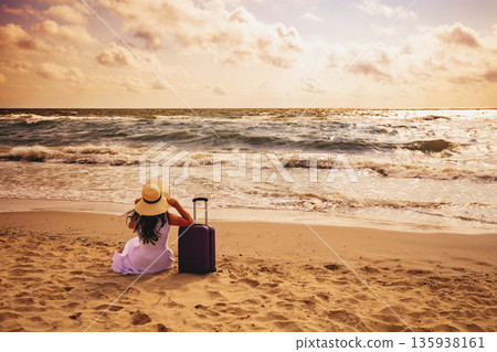 Seascape during sunrise. A woman sits on the beach in summer with a travel suitcase. The girl looks at the sea. Travel, happy holiday, summertime concept 135938161