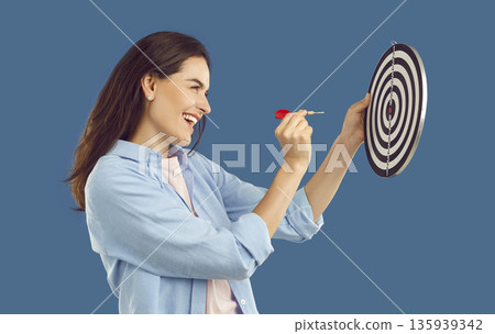 Happy brunette young woman in casual shirt aiming dart arrow at dartboard on blue background. 135939342