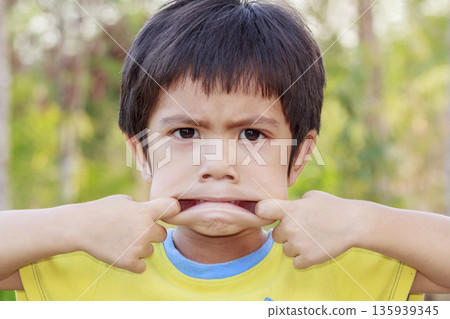 Portrait of Asian little boy doing a funny pose pulling the corner of his mouth with his fingers outdoor 135939345