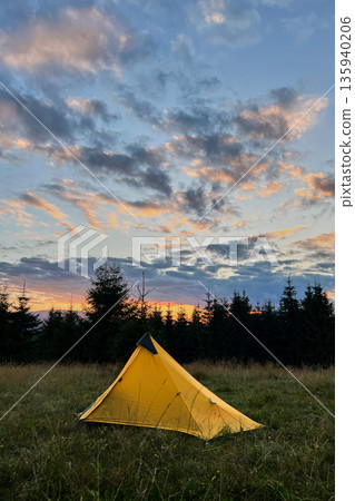 Yellow tourist tent set up in grassy field at sunset, surrounded by trees in evening. Sky painted with vibrant hues of orange, pink, and purple as sun sets, casting warm glow over serene campsite. Yellow tourist tent set up in grassy field at sunset, surrounded by trees in evening. Sky painted with vibrant hues of orange, pink, and purple as sun sets, casting warm glow over serene campsite. 135940206