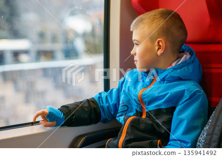 Thoughtful young boy in warm jacket sitting by train window, looking outside during winter travel. Concept of childhood, transportation, journey Thoughtful young boy in warm jacket sitting by train window, looking outside during winter travel. Concept of childhood, transportation, journey 135941492