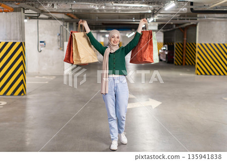 A happy woman wearing a hijab is carrying shopping bags in a parking garage, celebrating her purchases 135941838