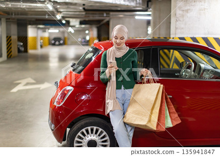 A woman in a hijab stands by her car in a parking garage, holding shopping bags and looking at her phone 135941847