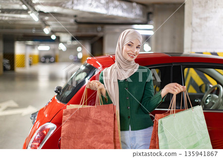 A smiling woman wearing a hijab stands next to a red car, holding shopping bags in a parking garage A smiling woman wearing a hijab stands next to a red car, holding shopping bags in a parking garage 135941867