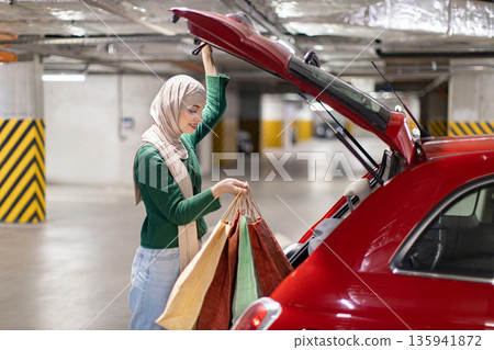 A woman wearing a hijab loads shopping bags into the trunk of her red car in a parking garage 135941872