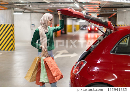 A woman wearing a hijab loads shopping bags into the trunk of her red car in a parking garage 135941885