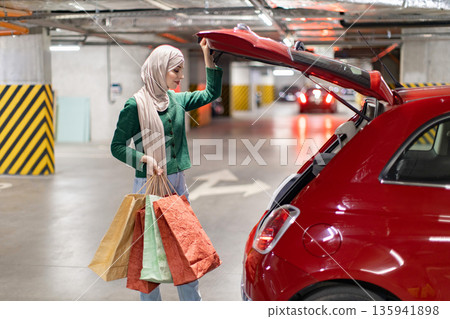 A woman wearing a hijab loads shopping bags into the trunk of her red car in a parking garage 135941898