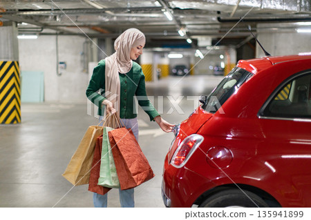 A woman wearing a hijab opens the trunk of her car after a shopping trip in a parking garage 135941899
