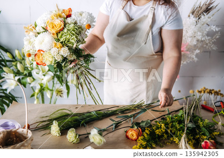 Hands of florist hold large floral bouquet over workbench in studio. Sustainable wedding decor, micro-wedding aesthetics, floral symbolism. Hands of florist hold large floral bouquet over workbench in studio. Sustainable wedding decor, micro-wedding aesthetics, floral symbolism. 135942305