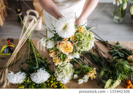Florist arranges fresh blossoms on paper covered table in bright studio. Artisan craftsmanship, small business growth, floral design. Florist arranges fresh blossoms on paper covered table in bright studio. Artisan craftsmanship, small business growth, floral design. 135942306