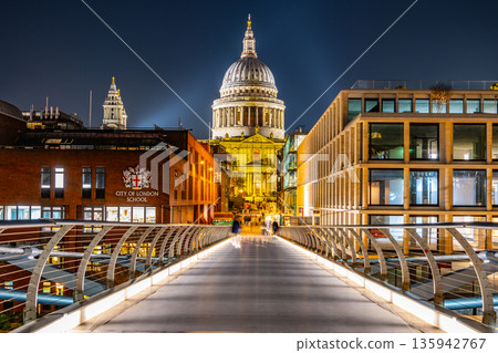 St Pauls Cathedral stands tall against the night sky. The scene captures the view from Millennium Bridge in London. People walk across the bridge enjoying the city lights and architecture. St Pauls Cathedral stands tall against the night sky. The scene captures the view from Millennium Bridge in London. People walk across the bridge enjoying the city lights and architecture. 135942767