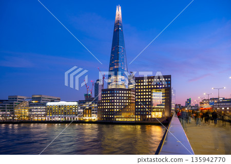 The Shard rises above the Thames River as day turns to night in London. People move along the walkway, enjoying the city as the lights begin to shine. This scene captures urban life. 135942770