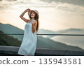 Charming woman in white dress posing at mountain sunset view, Beautiful female with straw hat against golden hour mountains background 135943581