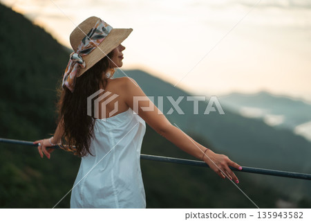 Charming woman in white dress posing at mountain sunset view, Beautiful female with straw hat against golden hour mountains background 135943582
