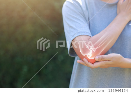 Close-up woman holding painful injured elbow with red inflamed spot and X-ray bone skeleton overlay. Medical concept of joint pain, fracture, arthritis, tendonitis, muscle injury, outdoor background. 135944154