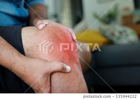 Close-up of hand holding painful injured knee with red inflamed spot and bruising. Medical concept of joint pain, arthritis, bone injury, muscle ache, or sprain in outdoor sunlight background. Close-up of hand holding painful injured knee with red inflamed spot and bruising. Medical concept of joint pain, arthritis, bone injury, muscle ache, or sprain in outdoor sunlight background. 135944222